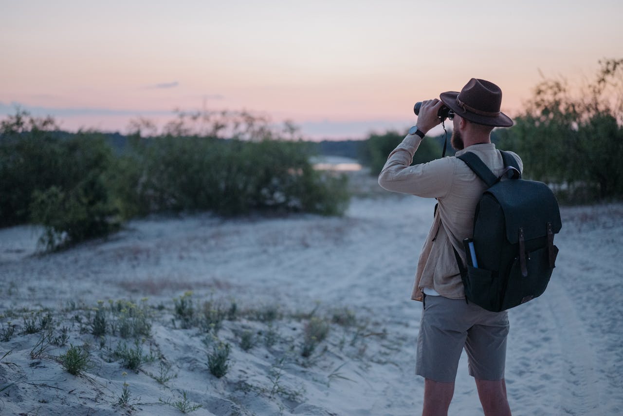 Offerings Man with backpack observing nature through binoculars during a scenic sunset.